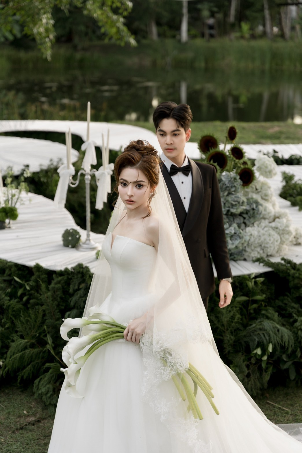 คู่บ่าวสาวในพิธีกลางแจ้ง ยืนอยู่ใต้ซุ้มดอกไม้